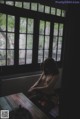 A naked woman sitting at a table in front of a window.